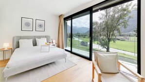A modern bedroom with a large bed, a bedside table, a chair, and a view of a green landscape through large windows. - An oasis of peace in nature (Cassiopea 13 Residenza Ticino) (Bellinzona)