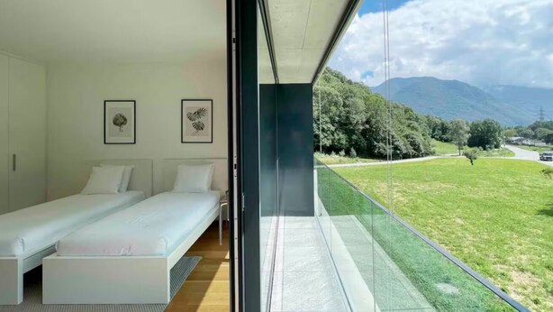 A modern bedroom with a large window overlooking a grassy landscape and mountains. - An oasis of peace in nature (Sirio 09 Residenza Ticino) (Bellinzona)