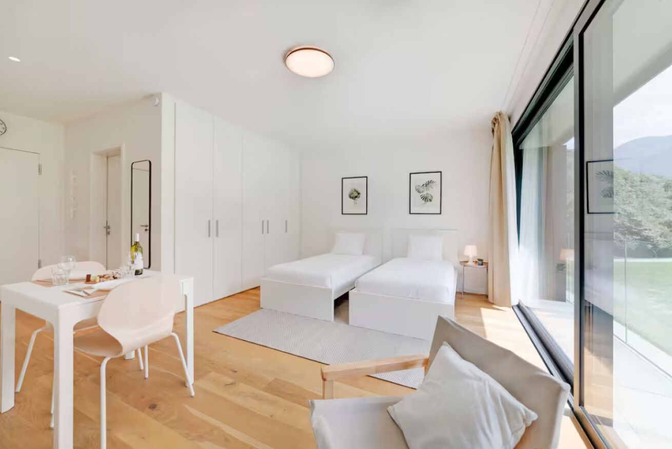 Ein modernes, minimalistisches Schlafzimmer mit Bett, Nachttischen und einem großen Fenster mit Blick ins Freie.