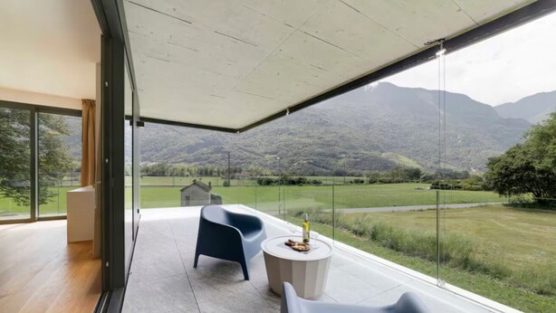 A modern room with a large glass window, a round table with a bottle, and two chairs. - An oasis of peace in nature (Hydra 06 Residenza Ticino) (Bellinzona)