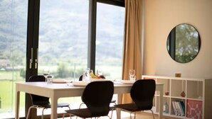 A dining area with a table set for two, large windows with a view of greenery, and a round mirror on the wall. - An oasis of peace in nature (Hydra 06 Residenza Ticino) (Bellinzona)
