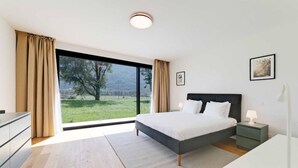 A modern bedroom with a large bed, a nightstand, a dresser, and a view of a green landscape through a large window. - An oasis of peace in nature (Talitha 05 Residenza Ticino) (Bellinzona)