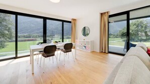 A modern room with a dining table, chairs, a white cabinet, and large windows offering a view of a green landscape and mountains.