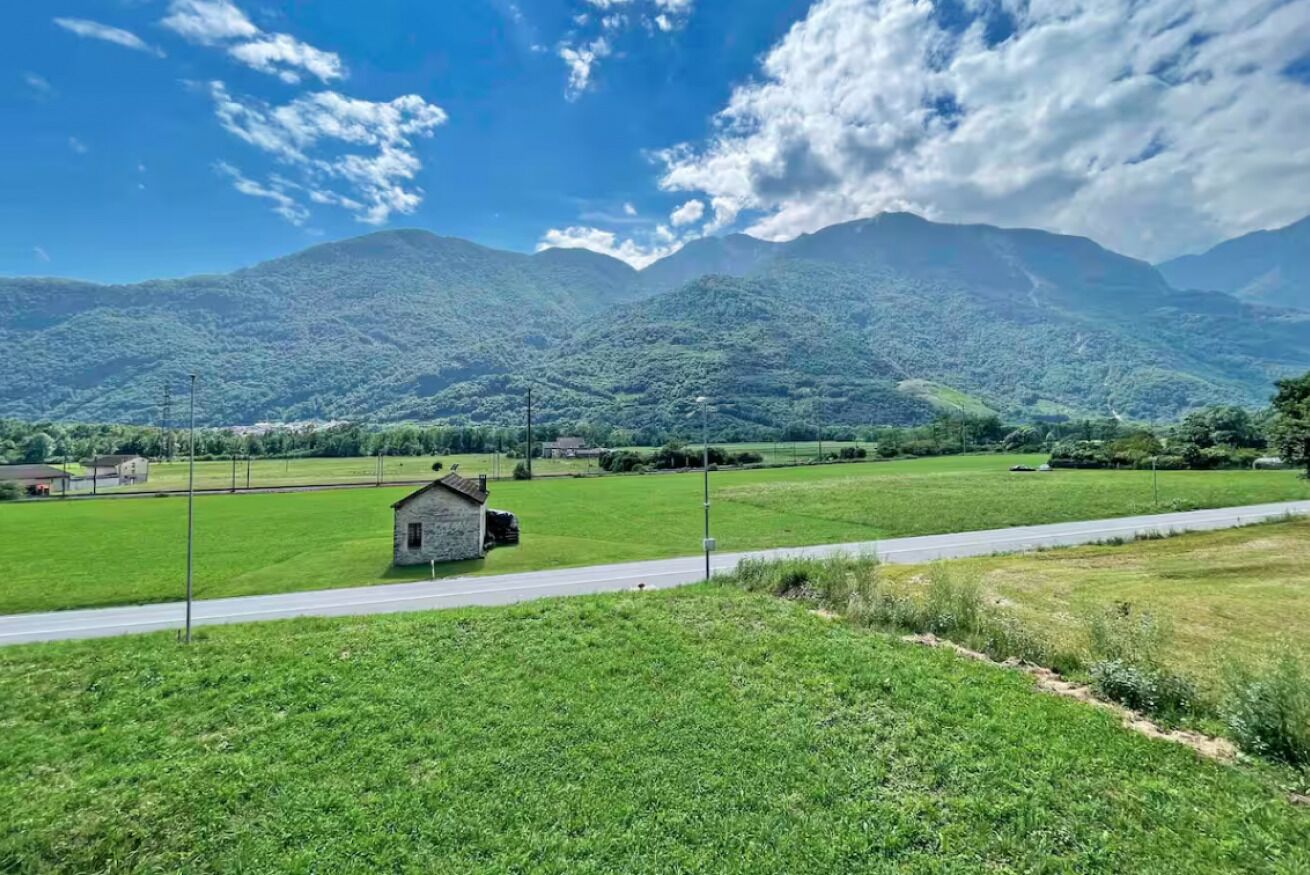A rural landscape with a small stone building near a road, surrounded by green fields and mountains in the background.