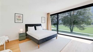 A modern bedroom with a large bed, a nightstand, a desk, and a view of a green landscape through large windows.