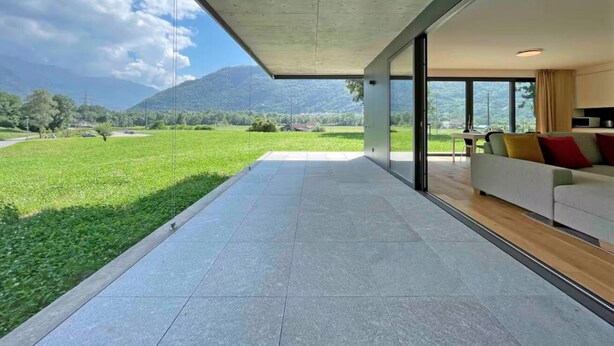 A modern living room with a large glass door leading to a balcony overlooking a green landscape and mountains.