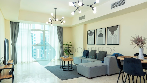 A modern living room with a grey sectional sofa, a dining table with chairs, a round coffee table, and a large window with curtains. - Bloomfields Cozy 1BR Ocean Terrace (Abu Dhabi)