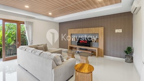 A modern living room with a sofa, a glass coffee table, and a flat-screen TV mounted on a wooden panel. - Amaya Villa by Kozystay - Canggu (Canggu)