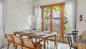 A dining area with a wooden table set for four, surrounded by wicker chairs. There are two pendant lights above the table, and a fire extinguisher mounted on the wall. - Amaya Villa by Kozystay - Canggu (Canggu)