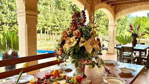 Un espace de restauration en plein air avec une table en bois dressée pour un repas, entouré d’arcades et offrant une vue sur une piscine et la verdure.