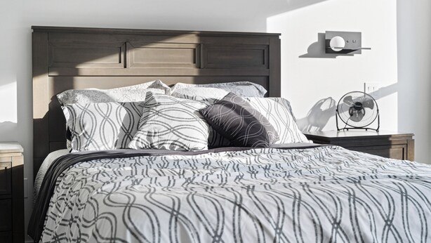 A neatly made bed with patterned bedding, a wooden headboard, and a bedside table with a fan.