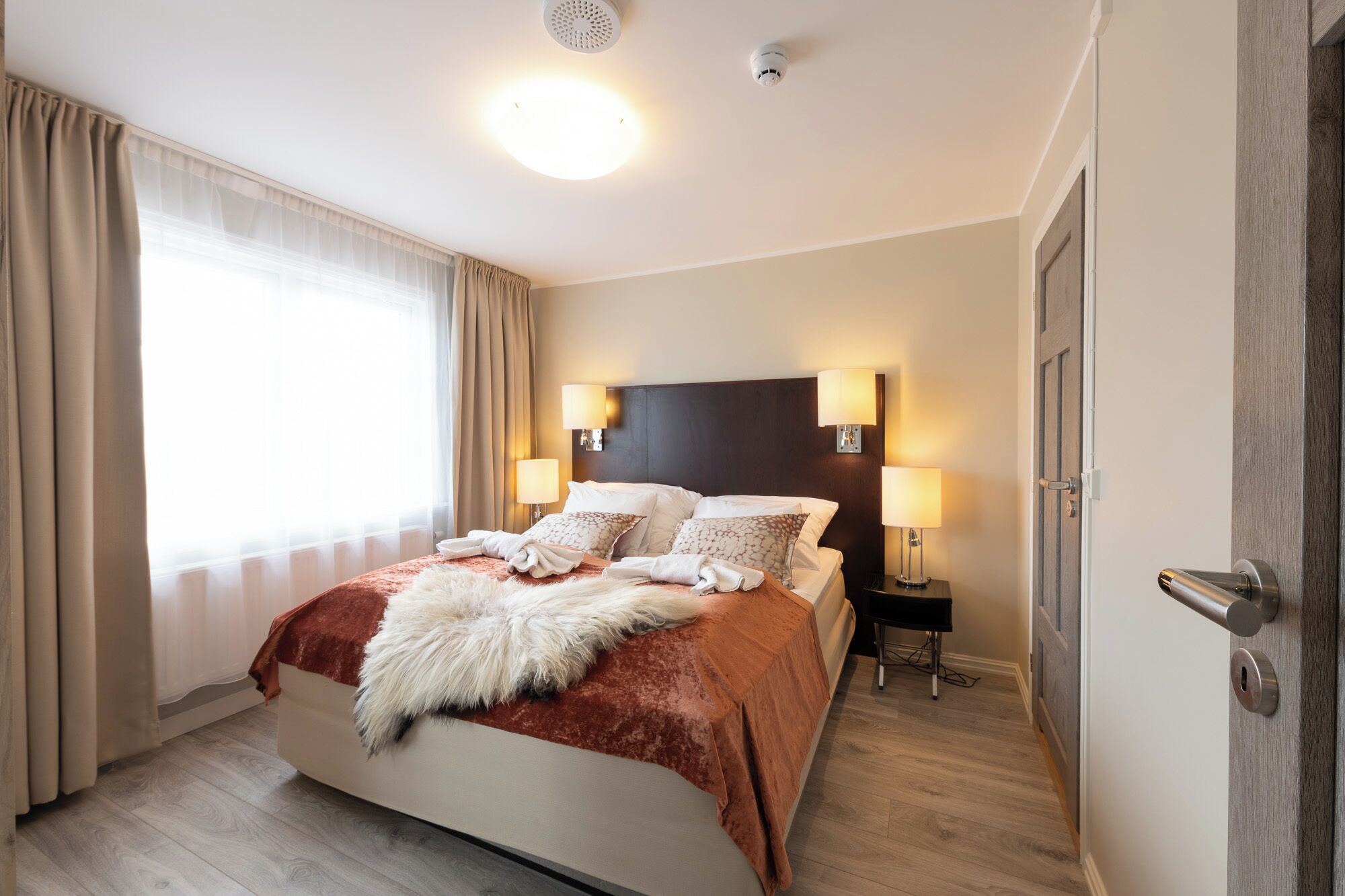 Une chambre d’hôtel avec un grand lit, deux lampes de chevet, une tête de lit en bois et une porte dotée d’une poignée moderne.