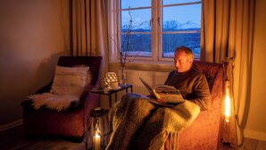 A cozy room with a person reading a book by a window. There's a chair with a blanket, a small table with a candle, and a vase with branches.