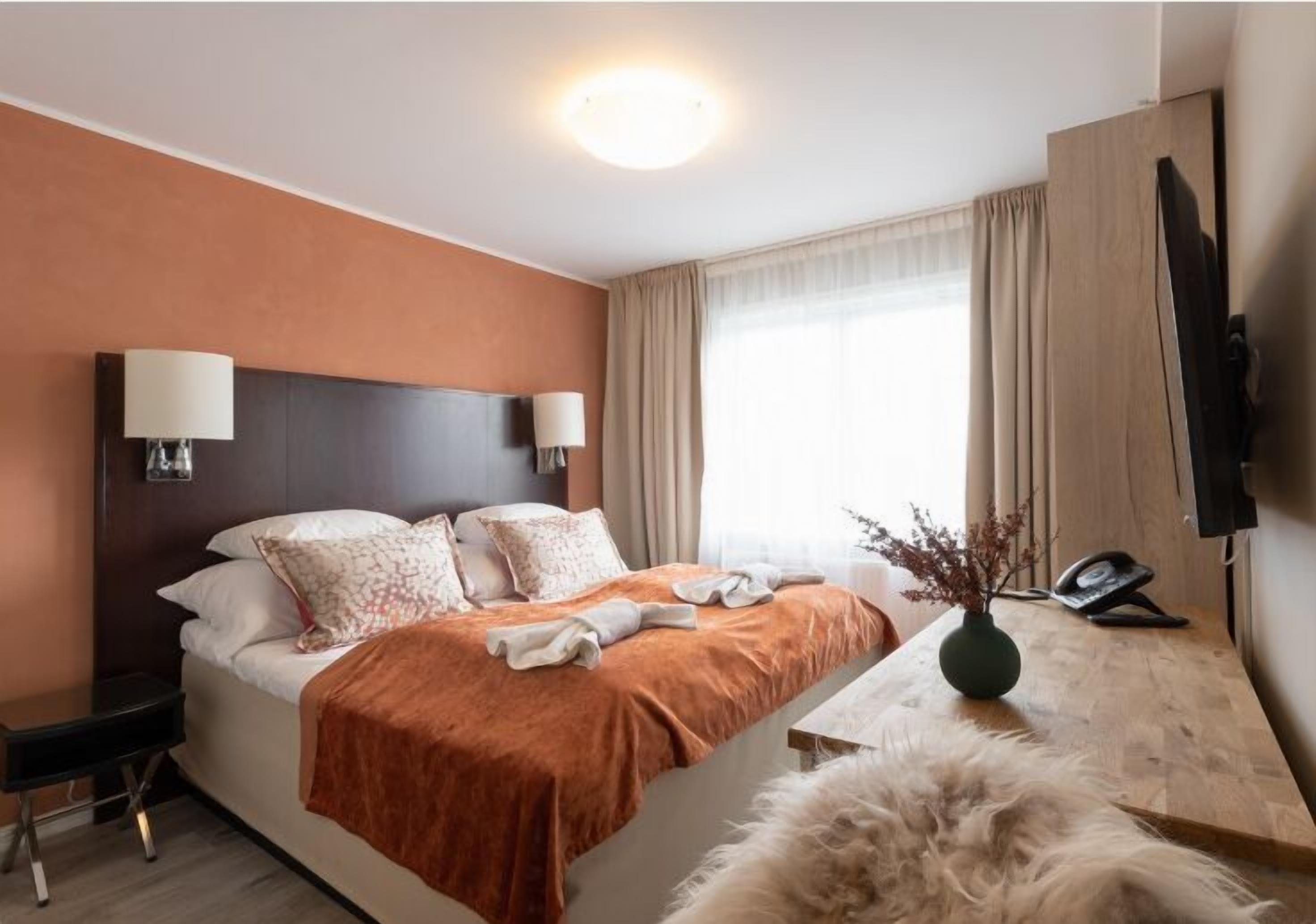 A hotel room with a bed, bedside lamps, a desk with a telephone, a vase with dried branches, and a television on the wall.