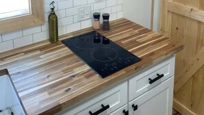 A kitchen with wooden shelves holding dishes and glasses, a black induction cooktop, and a window. - Mountain Top Tiny House by Beyond Expectations: 1BR w/ Mountain Views (Sevierville)