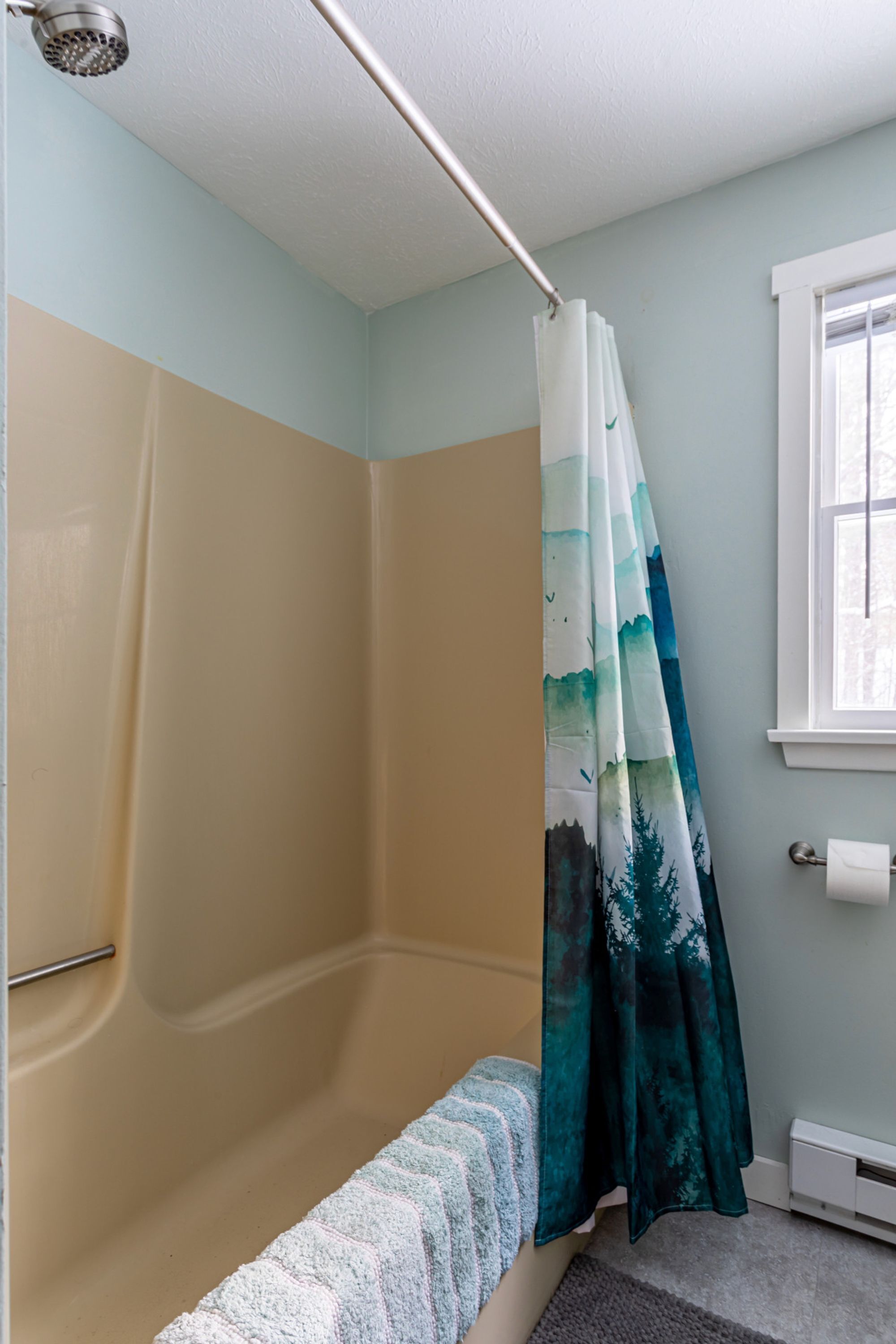 A bathroom with a bathtub, a shower curtain with a forest design, and a window with a view of trees.