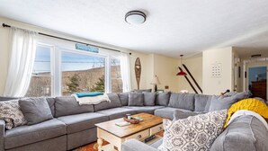 A modern living room with a grey sectional sofa, a wooden coffee table, and a view of a mountain landscape through large windows. - Ski Hike Lodge 5BR w Fire Pit Cozy Spaces (Maplecrest)