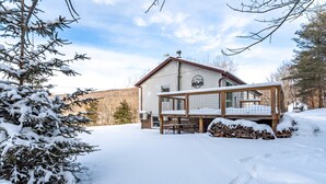 A snow-covered lodge with a wooden deck and a large window overlooking a snowy landscape. - Ski Hike Lodge 5BR w Fire Pit Cozy Spaces (Maplecrest)