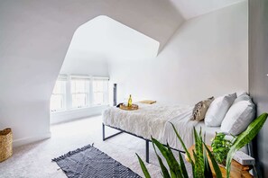 Un dormitorio con techo inclinado, una cama con ropa de cama blanca, una mesita de noche con lámpara y un ventanal grande.