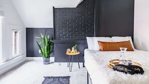 Ein modernes Schlafzimmer mit einem weißen Bett, einer Wand aus schwarzen Ziegelsteinen und einem kleinen Nachttisch mit einer Pflanze.