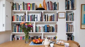 Een eetruimte met een houten tafel, stoelen, een vaas met bloemen, een schaal met fruit en een theepot.