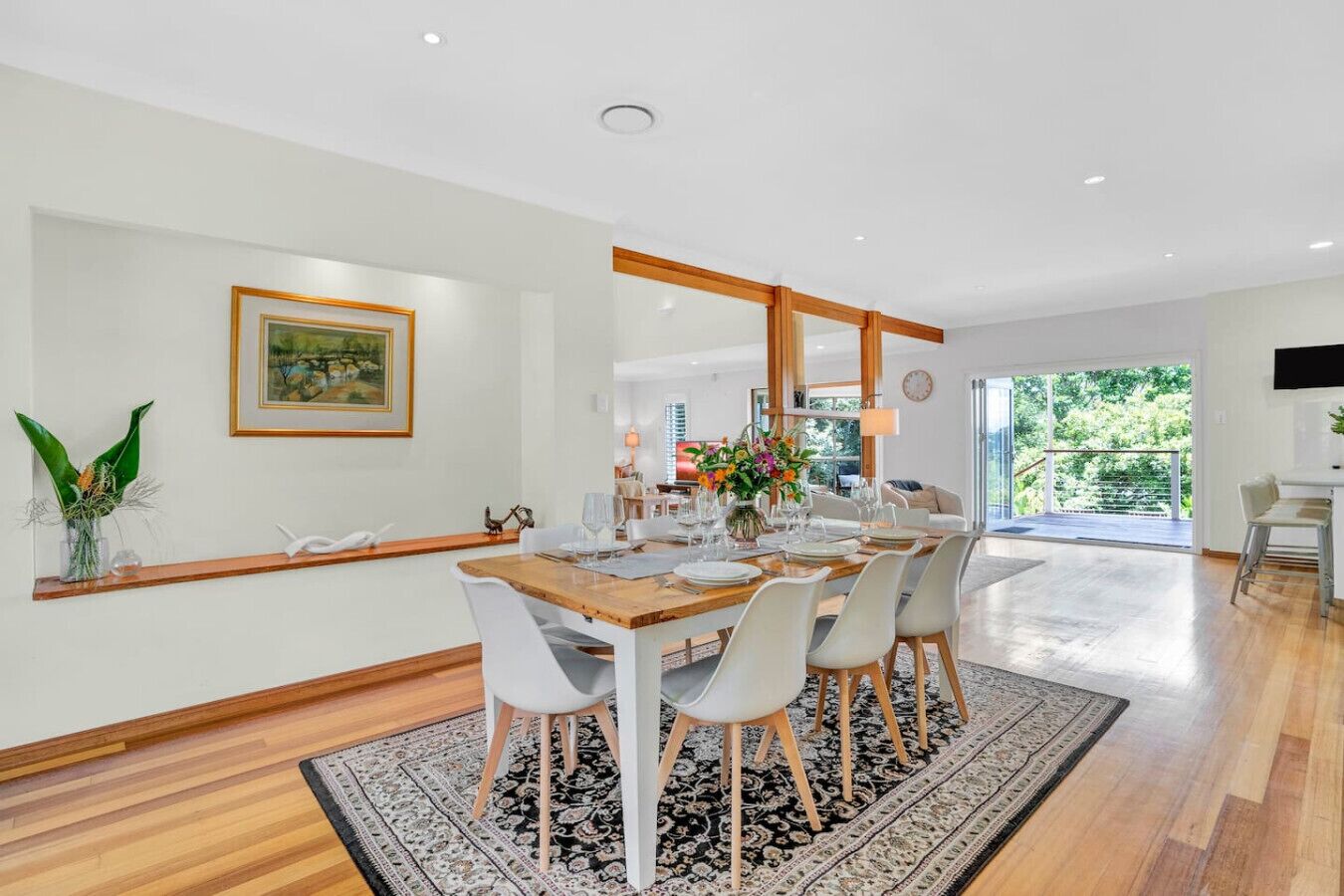 A modern dining area with a wooden table set for six, surrounded by white chairs. A large mirror hangs above the table, and a framed painting is on the wall to the left.