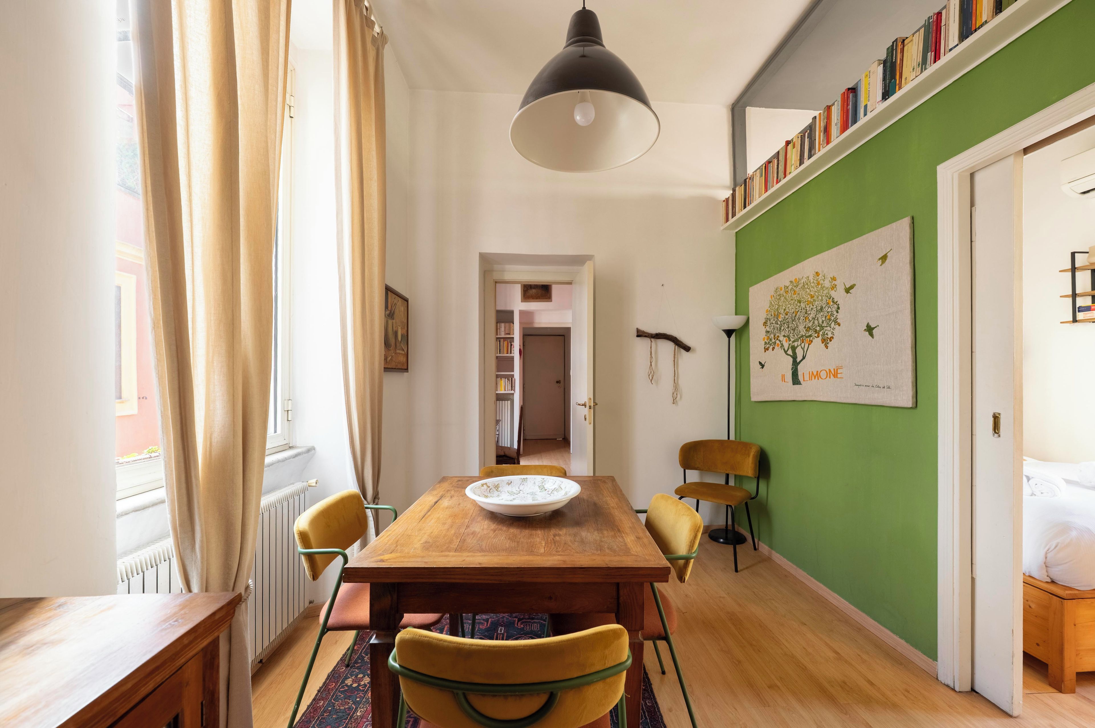 A dining area with a wooden table, chairs, a hanging light fixture, and a green wall with a framed picture.