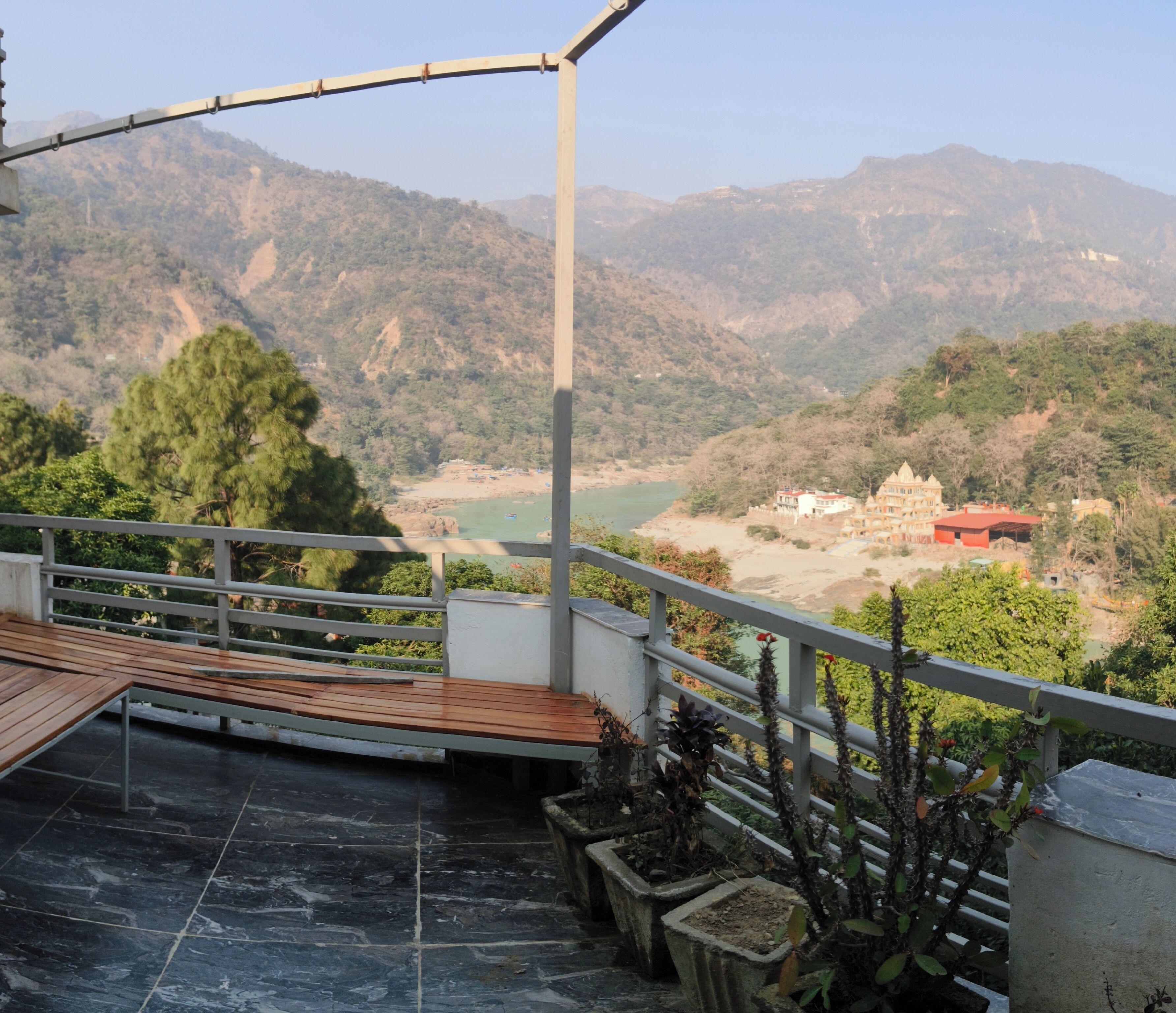 A balcony with a wooden floor and metal railing, overlooking a mountainous landscape with a river and buildings.
