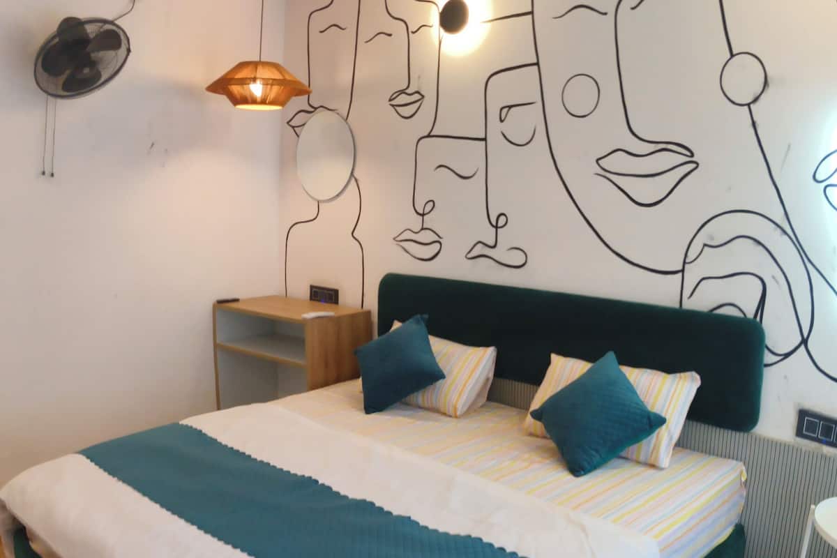 A bedroom with a bed, a wall-mounted fan, a shelf, and a wall art piece.