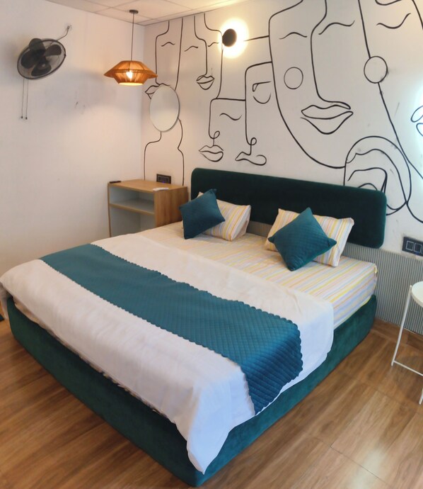 A bedroom with a bed, a wall-mounted fan, a shelf, and a wall art piece. - Art Bliss Hostel (Rishikesh)