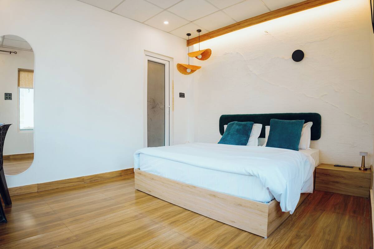 A modern bedroom with a wooden floor, a bed with white bedding and teal pillows, a wooden nightstand, and a wall-mounted light fixture.