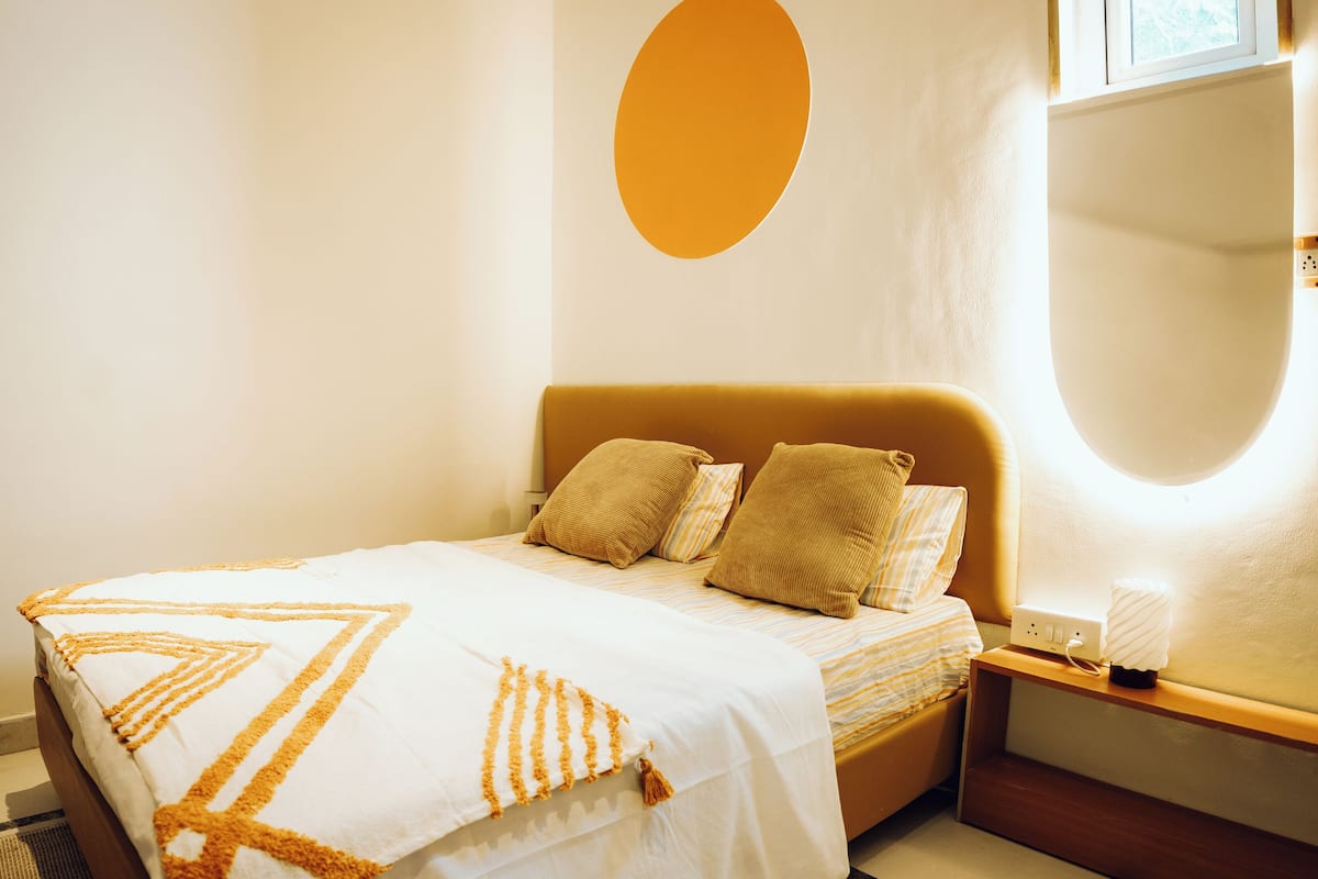 A neatly made bed with a striped bedspread and two pillows, a bedside table with a lamp, and a wall-mounted mirror.