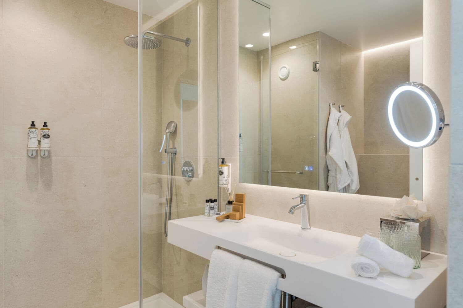 Une salle de bain moderne avec une douche à parois de verre, un grand miroir avec un éclairage rond et un meuble-lavabo blanc doté d’un porte-serviettes.