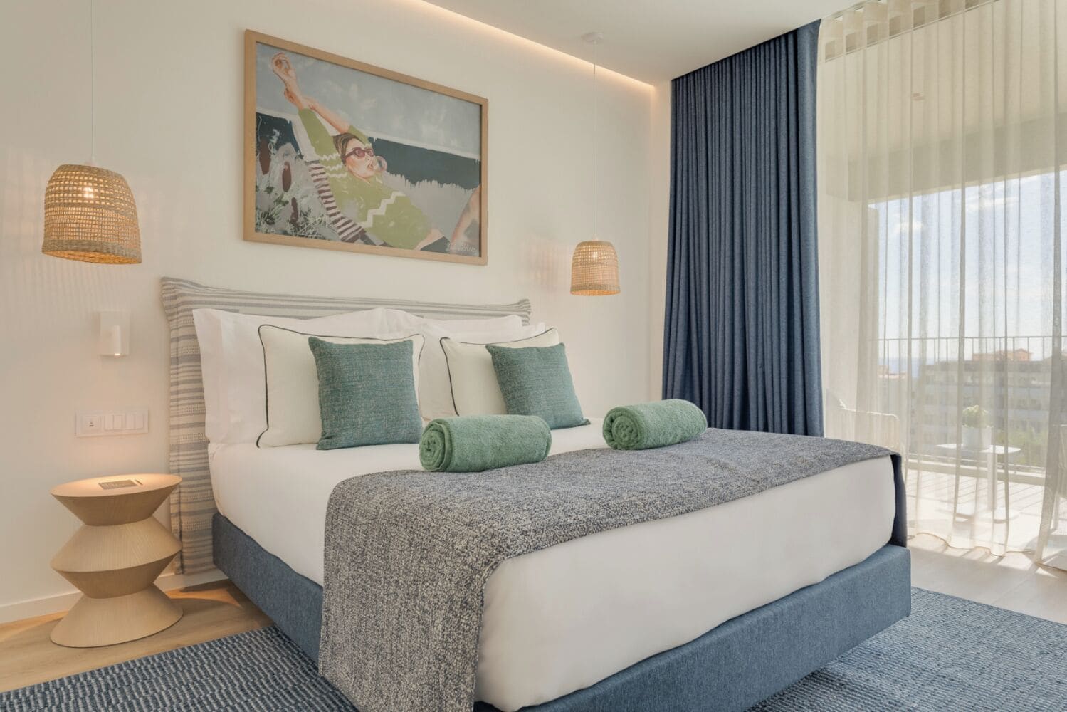 A modern hotel room with a large bed, two bedside lamps, a painting on the wall, and a view through sheer curtains.