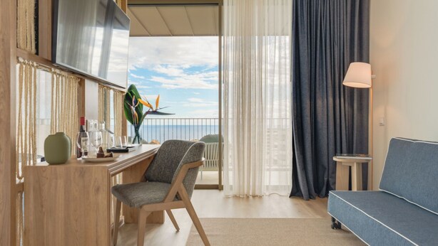 A modern hotel room with a wooden desk, a blue sofa, and a balcony with a view of the sky and buildings.