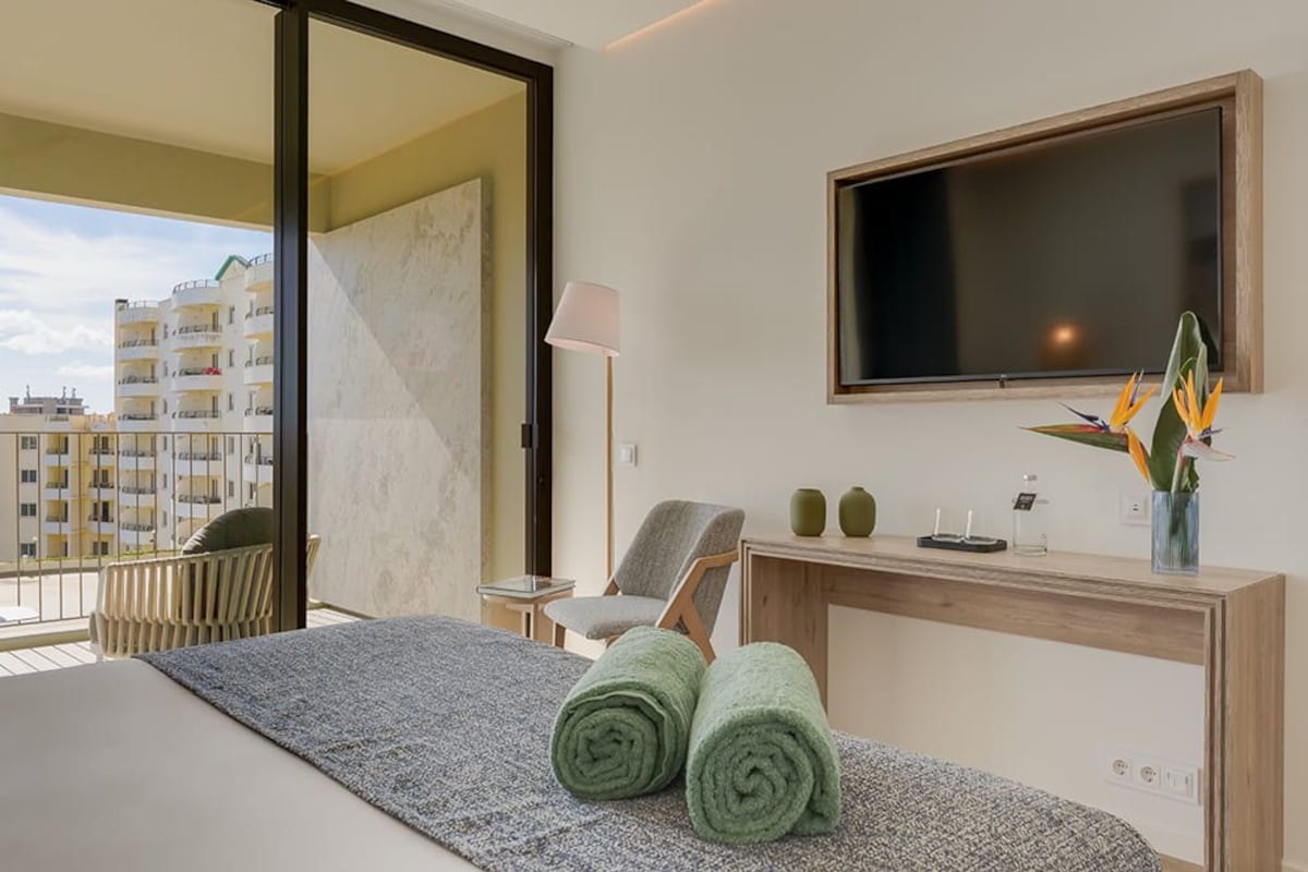 A hotel room with a bed, a desk with a lamp, a chair, a TV, and a view of buildings.