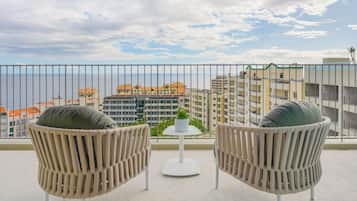 Un balcón con dos sillas de mimbre y una mesita, con vistas a un paisaje urbano con edificios y cielo despejado.