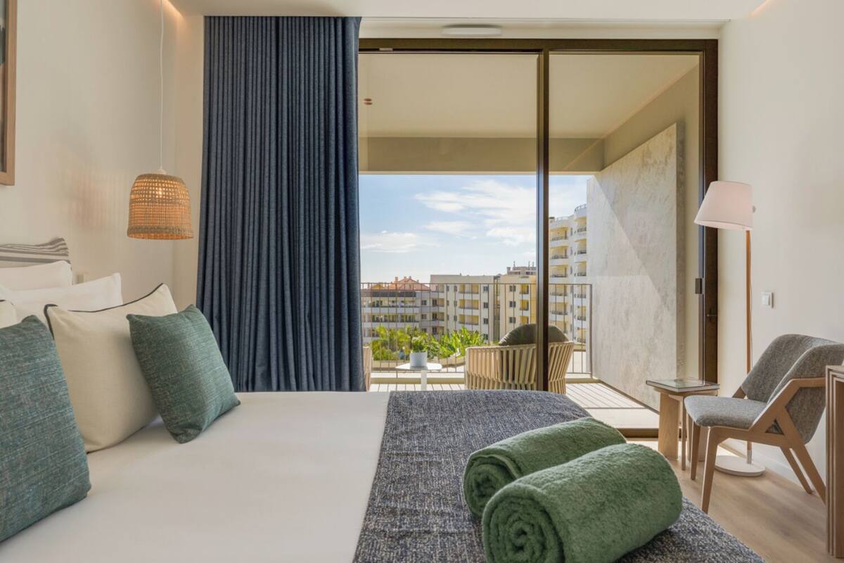 Ein Hotelzimmer mit einem großen Bett, einem Balkon mit Blick auf Gebäude, einem kleinen Tisch mit Stühlen und einer Lampe.