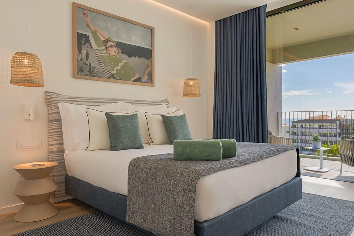 Une chambre moderne avec un grand lit, un tableau, deux lampes fixées au mur et une vue sur la ville depuis un balcon.