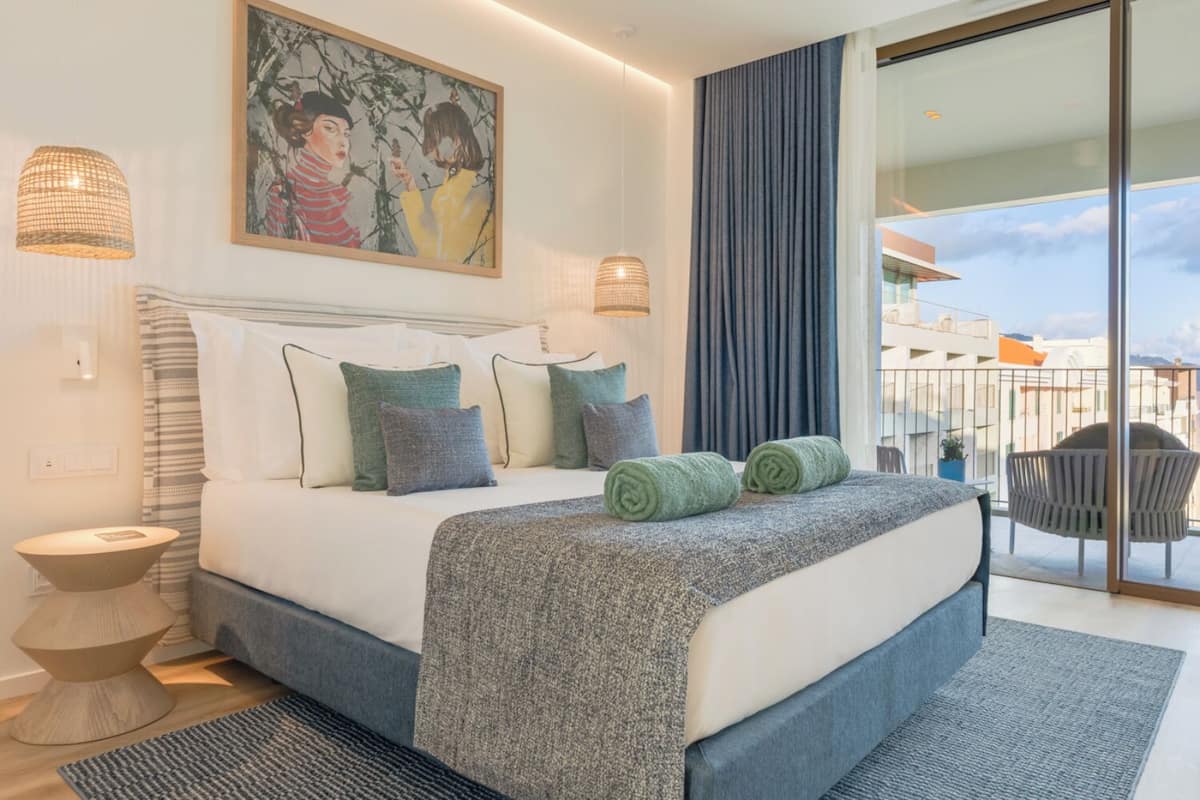 Ein modernes Schlafzimmer mit einem großen Bett, einem Gemälde darüber, einem Balkon mit Blick und einem Stuhl davor.