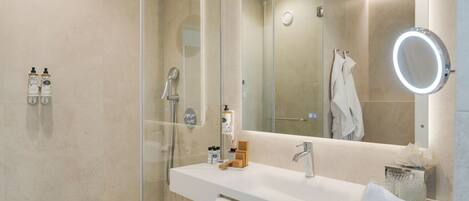 A modern bathroom with a glass-enclosed shower, a large mirror with a round light, and a white vanity with toiletries.