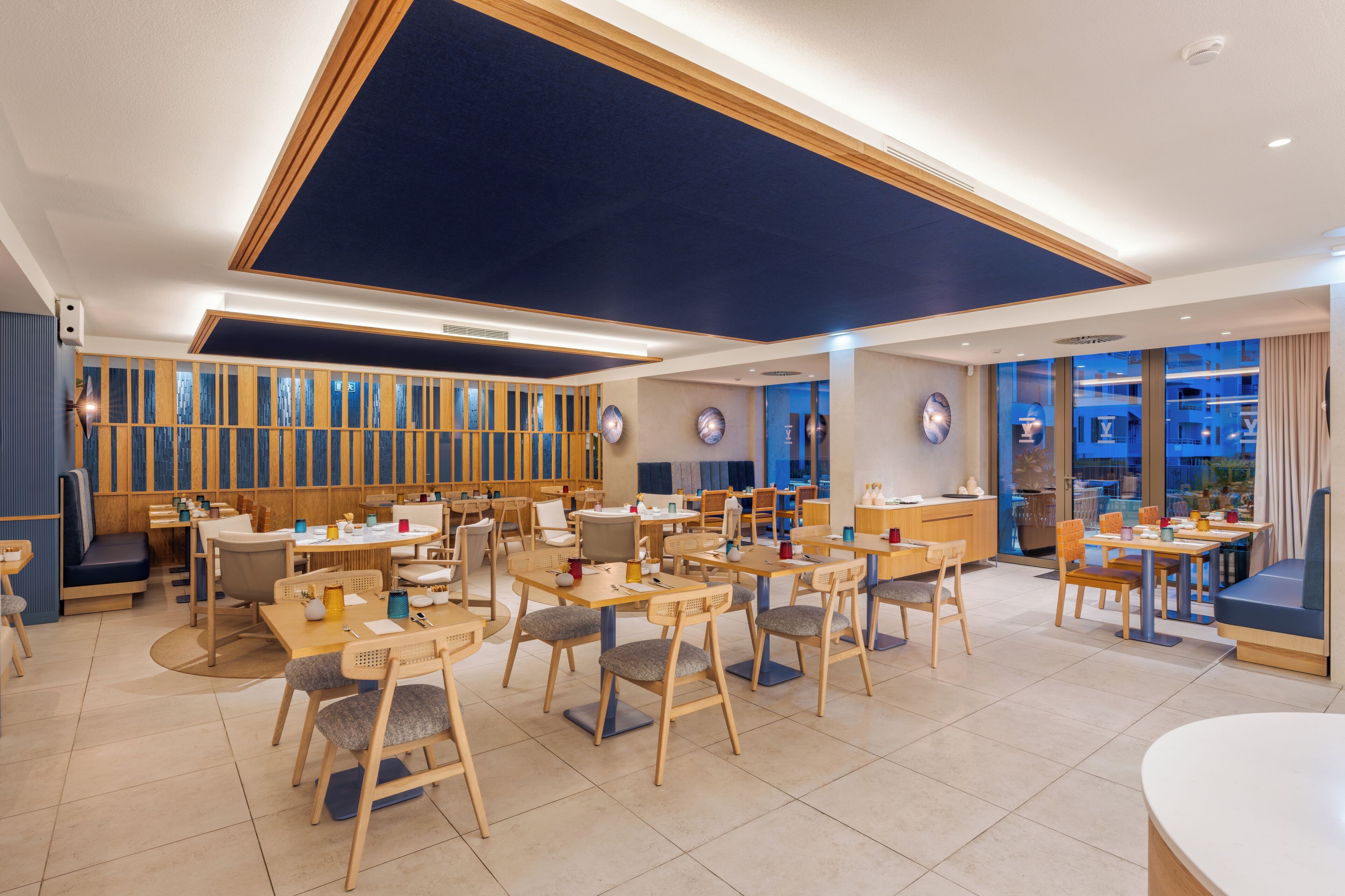 A modern dining area with multiple tables and chairs, a high ceiling with recessed lighting, and a view through the windows.