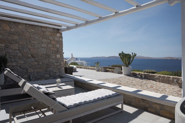 A sunlit outdoor lounge area with wicker lounge chairs and a stone wall backdrop, overlooking a coastal landscape. - Spacious 6-bedroom house with WiFi and AC in lovely Mykonos (Mykonos)