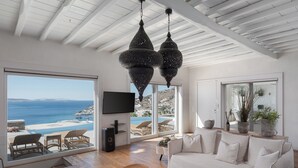 Uma sala de estar moderna com uma grande janela com vista para uma praia, um sofá branco e uma mesa de centro de madeira.