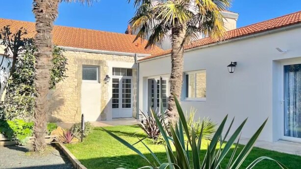 Un edificio a due piani con facciata in pietra, tetto in tegole rosse e un piccolo giardino con palme e prato.
