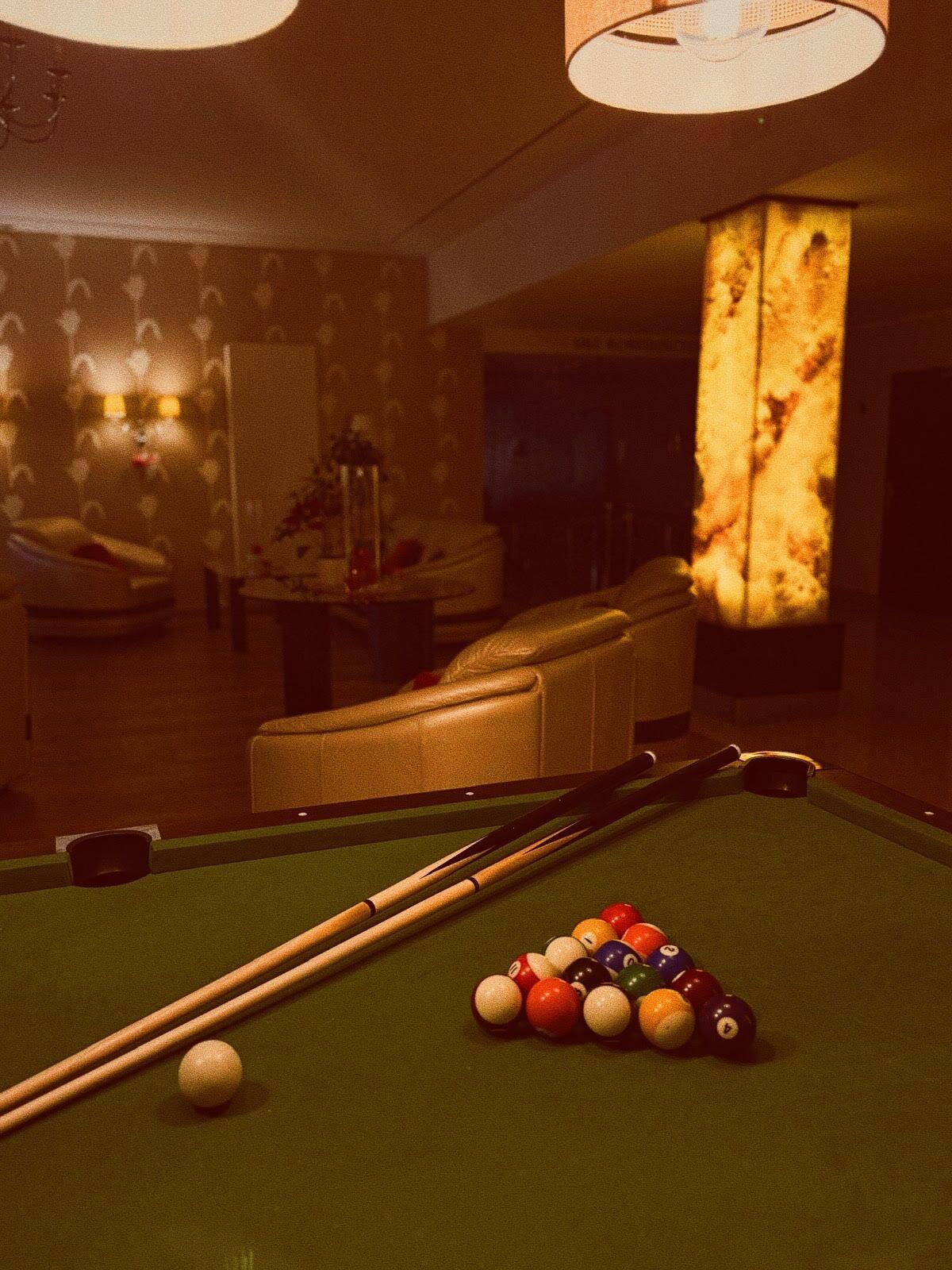 Ein Billardtisch mit Kugeln und Spielstöcken, ein Ledersofa und eine Lampe in einem schummrigen Raum.
