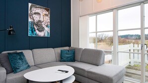 Un salon moderne avec un canapé gris, des coussins turquoise, une table basse blanche et une grande fenêtre donnant sur une clôture blanche et des arbres.