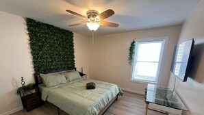 Un dormitorio con cama, ventilador de techo, mesita de noche con planta, ventana y televisor montado en la pared.