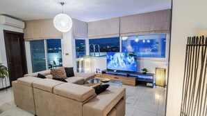 A modern living room with a large flat-screen TV, beige sofa, and a city view through large windows. - D Pool Top (L-Imsida)