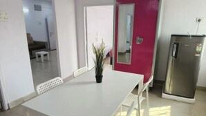 Un coin repas avec une table et des chaises blanches, une plante en pot, une porte rouge et un réfrigérateur.